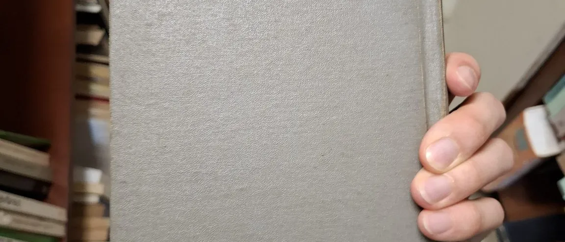A hand holding up a grey hardcover book inside a bookshop.