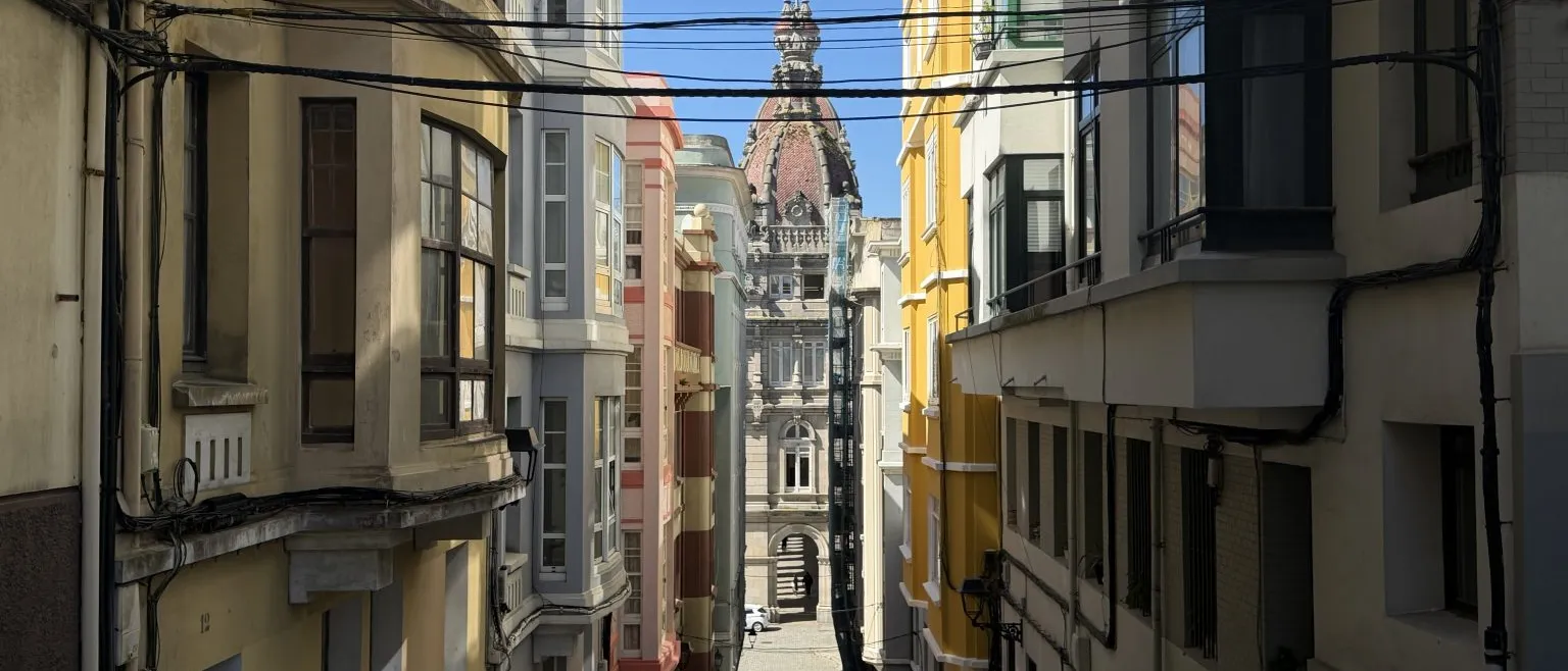 A narrow uphill street of colourful houses leading to a city hall with a reddish dome, under a clear blue sky.